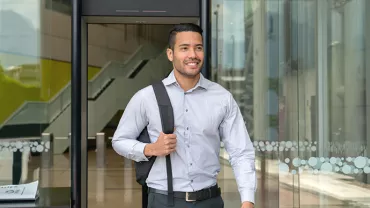 Smiling man with a backpack exits a glass building entrance.