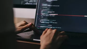 Person typing code on a laptop in a dimly lit room.