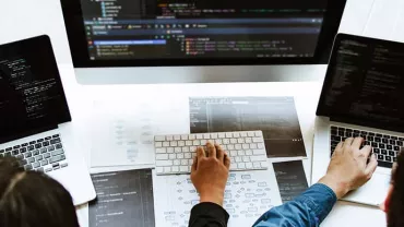 Three people collaborate on laptops with code on screens at a desk.