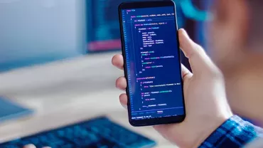 Person holding a smartphone displaying code, with a computer keyboard in the background.