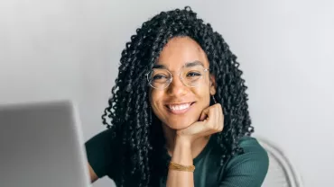 Woman with glasses smiling at laptop, hand on chin.