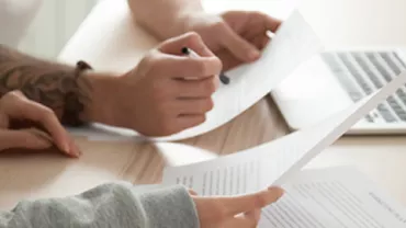 Two people reviewing documents at a desk with a laptop.