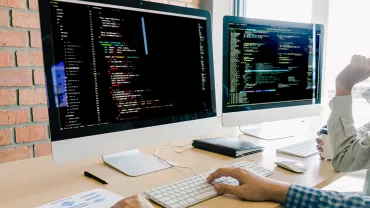 Two computer monitors displaying code in an office setting with two people working at a desk.