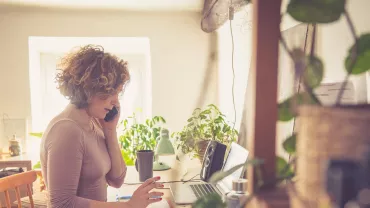 Woman working at home office desk with phone and laptop.