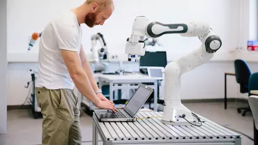 Engineer working on a laptop next to a robotic arm in a lab.