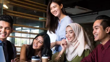 Five people gathered around a laptop, smiling and holding coffee, in an office setting.