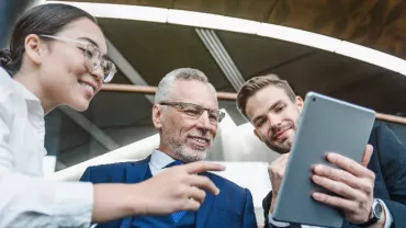 Three white-collar professionals looking at the screen of a tablet, all smiling