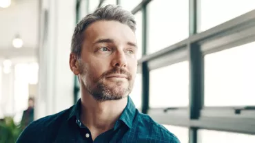 Man with a beard looking out a large window, wearing a blue shirt.