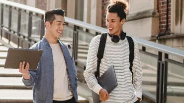 Two students with laptops walking and talking outside.