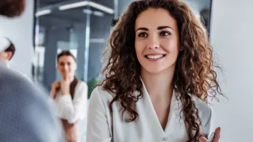 Person with curly hair smiling in office setting, two colleagues in the background.