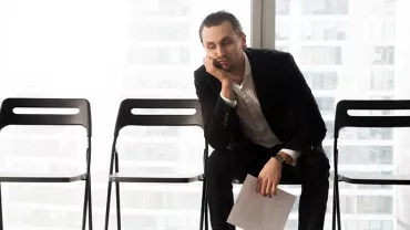 Man in a suit sitting on a chair, resting his head on his hand, looking tired, with empty chairs beside him.