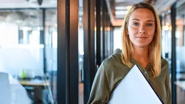 Woman standing in a modern office corridor holding a laptop.