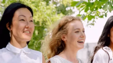 Three women smiling and walking outdoors under leafy trees.