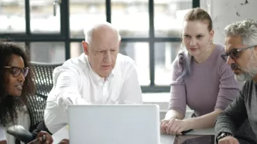 Group of four people collaborating around a laptop in a bright room.