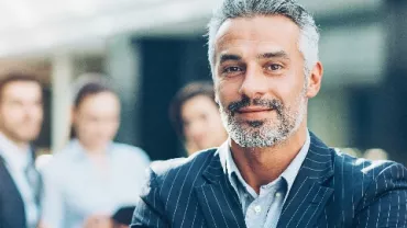 Smiling man in a business suit standing confidently in an office with blurred colleagues in the background.