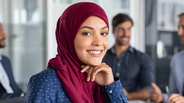 A female professional smiling, on a office background with three people dressed in corporate wear seated behind her