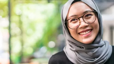 Woman in glasses and hijab smiling in an indoor setting.