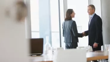A man and woman shaking hands in a conference room, indicating a formal agreement or partnership.