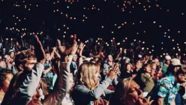 A large crowd cheering at a concert, with lights in the background.