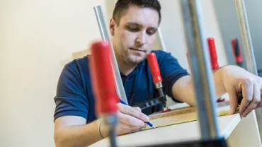 Man measuring and marking a wooden board using a pencil and clamps.