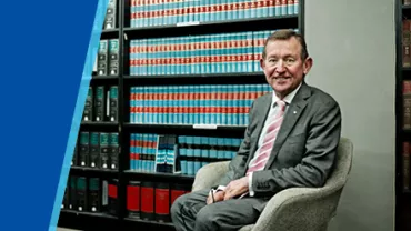 A man in a suit sits in a chair next to shelves filled with books.