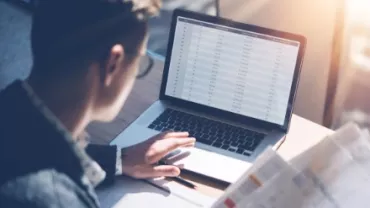 Person analyzing data on a laptop with spreadsheets and documents in hand.