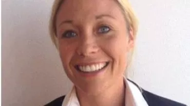 A smiling woman with blonde hair in a suit against a plain background.