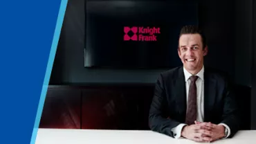 Businessman in suit smiling at desk with Knight Frank logo on screen behind.