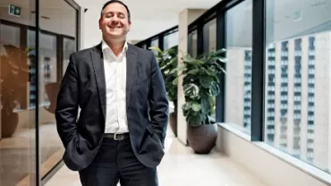 Smiling man in a suit standing in a bright office hallway.