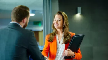 Woman in orange blazer shakes hands, holding a folder.