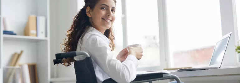 Woman in wheelchair smiling and holding a mug, sitting at a desk with a laptop.