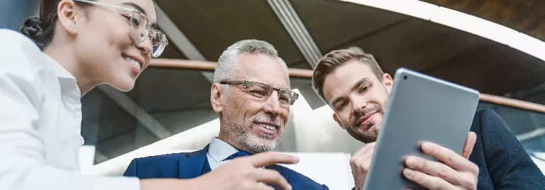 Three white-collar professionals looking at the screen of a tablet, all smiling