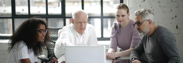 A group of office workers of different age groups and ethnicities looking at a computer screen at the same desk