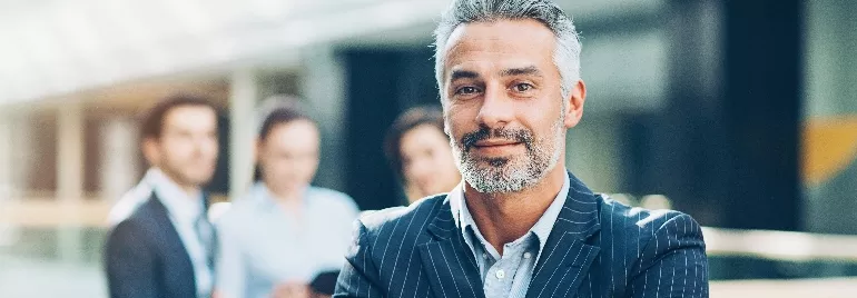 Smiling man in a business suit standing confidently in an office with blurred colleagues in the background.