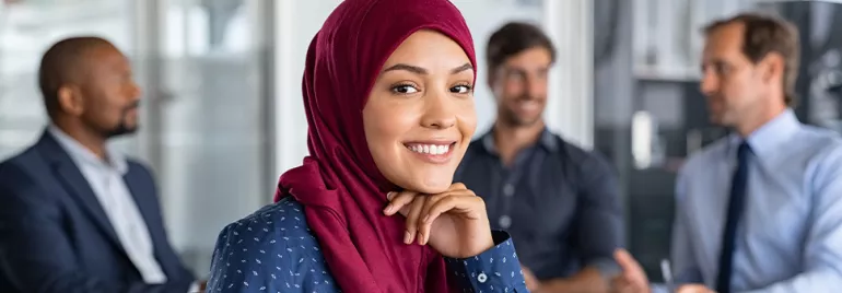 A female professional smiling, on a office background with three people dressed in corporate wear seated behind her