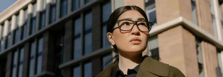 Female person with glasses stands in front of a modern building.